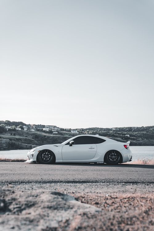 Free A modern white sports car parked by a scenic lakeside road under a clear sky. Stock Photo