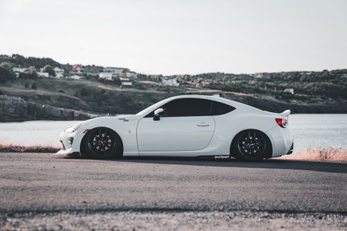 Free White sports car parked on a scenic coastal road with a backdrop of houses and greenery. Stock Photo