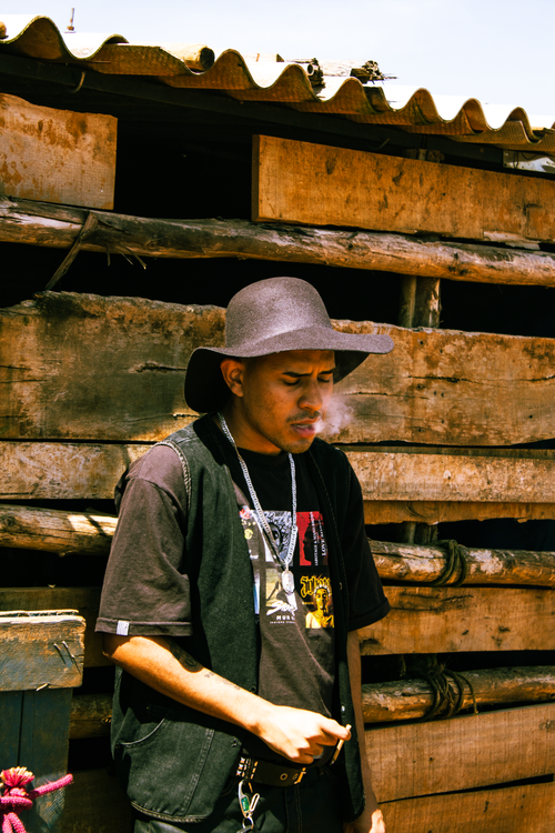 Free A young man with a hat smokes outside a rustic wooden structure on a sunny day. Stock Photo