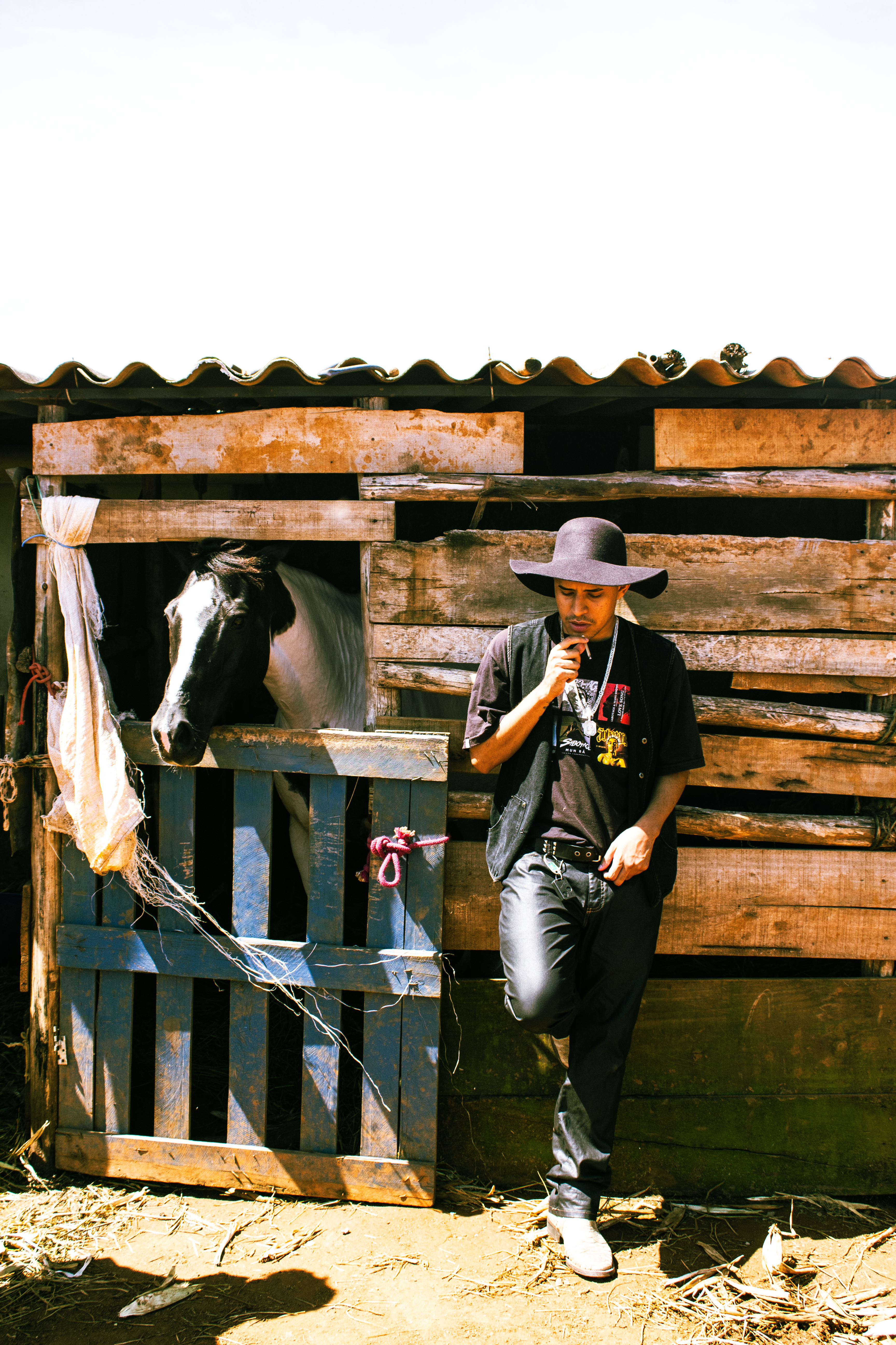 Man dressed in cowboy style standing by a rustic stable with a horse, captured in bright daylight.