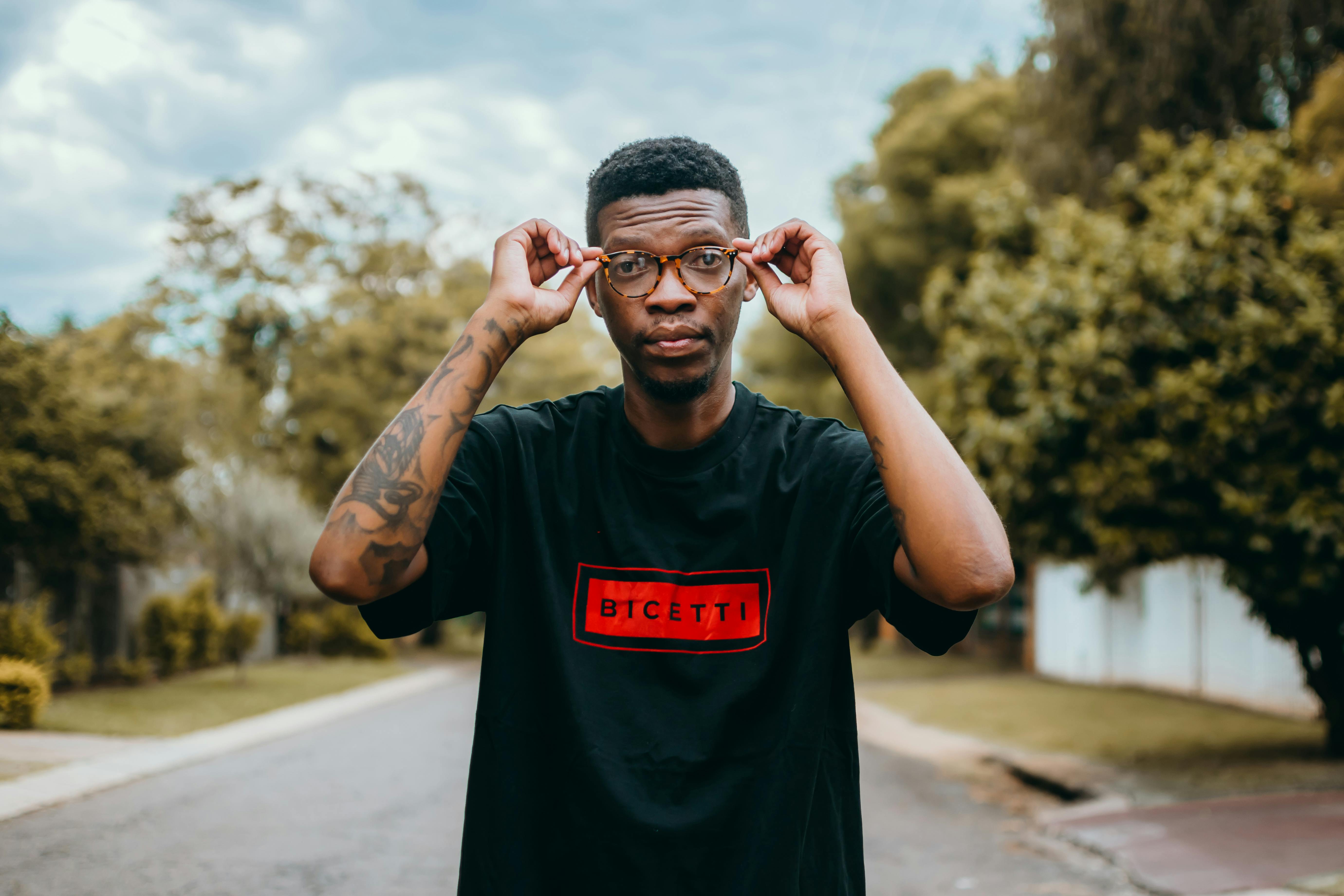 Free Portrait of a man adjusting glasses in Pretoria street with tattoo and casual attire. Stock Photo