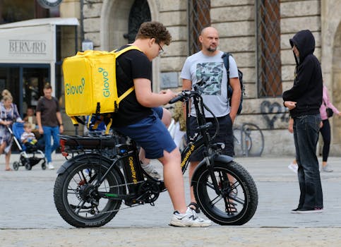 Courier with Glovo backpack on electric bike in a bustling city square, engaging with pedestrians.