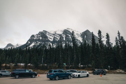 Snow-capped mountains and forest in Banff National Park with parked cars in foreground.