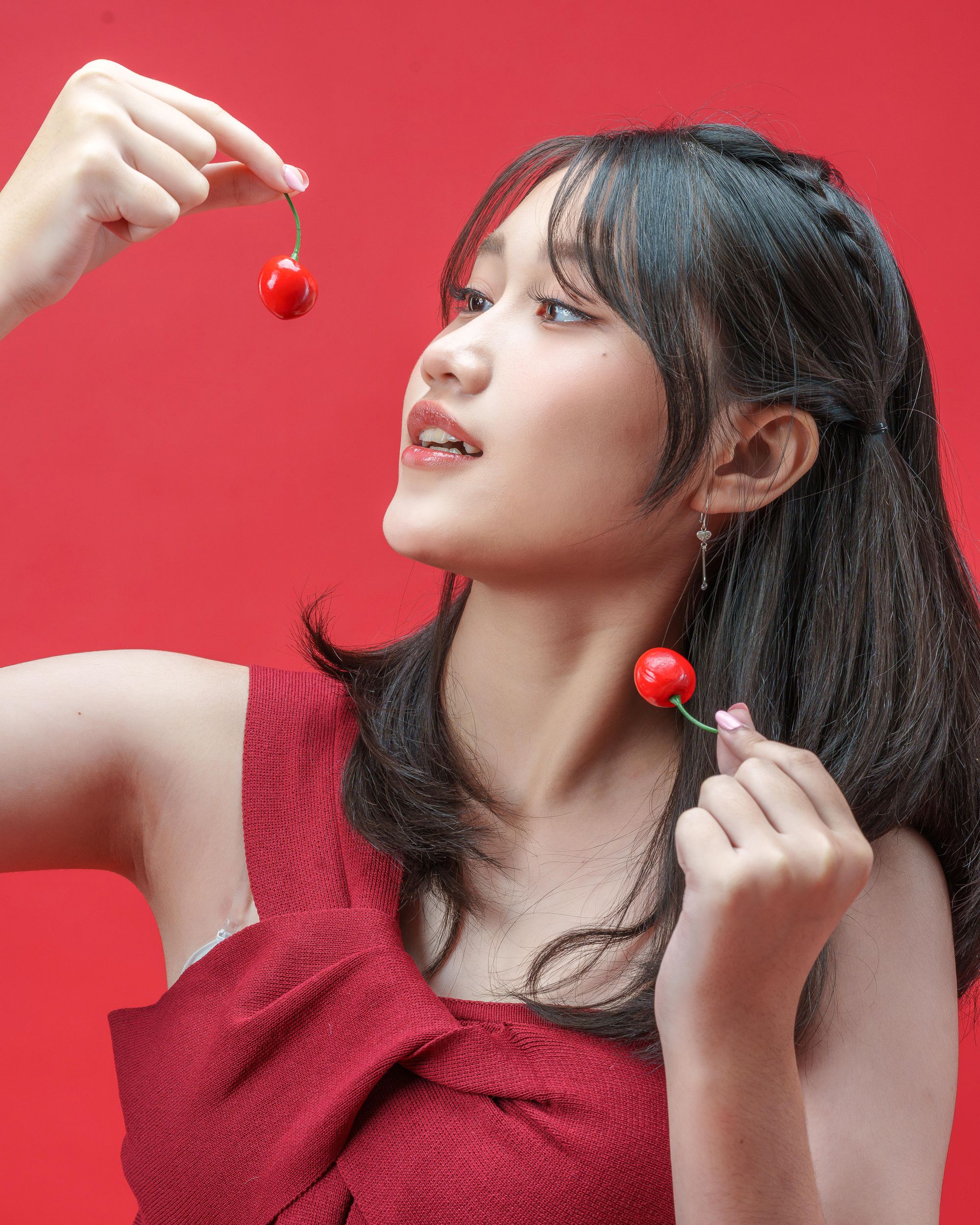 Portrait of a young woman in red holding cherries, vibrant red background.