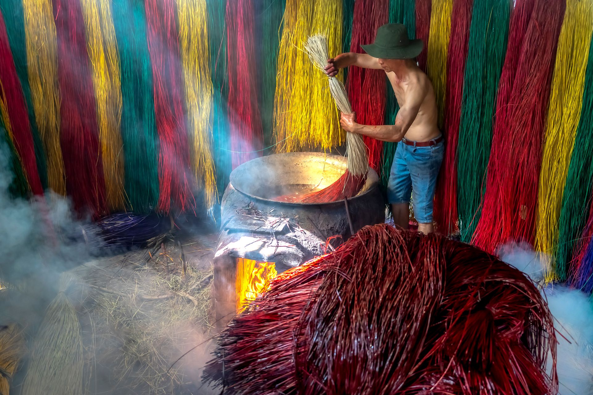 A man in a hat working in a vibrant, smoky workshop dyeing colorful fibers in a large pot.