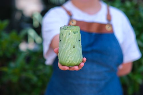 Free A barista holds out a glass of iced matcha latte with a blurred garden background. Stock Photo