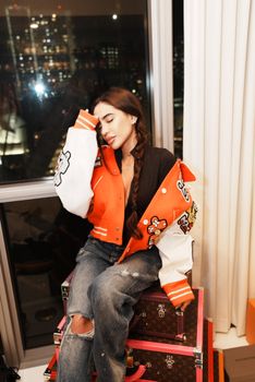 Young woman in a fashionable jacket posing indoors at night with city lights.