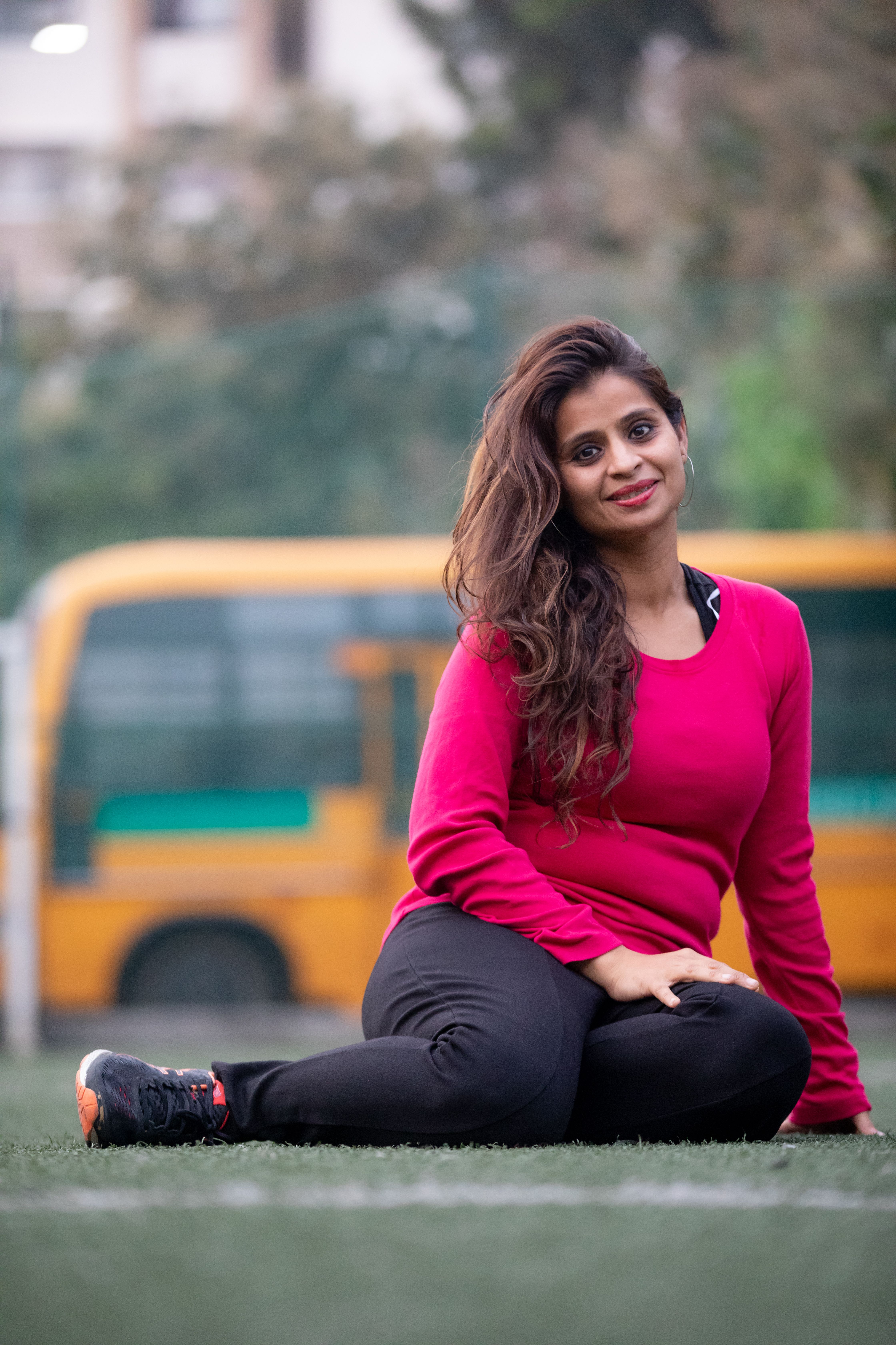 Smiling woman in pink sweater sitting on grass with blurred bus background.