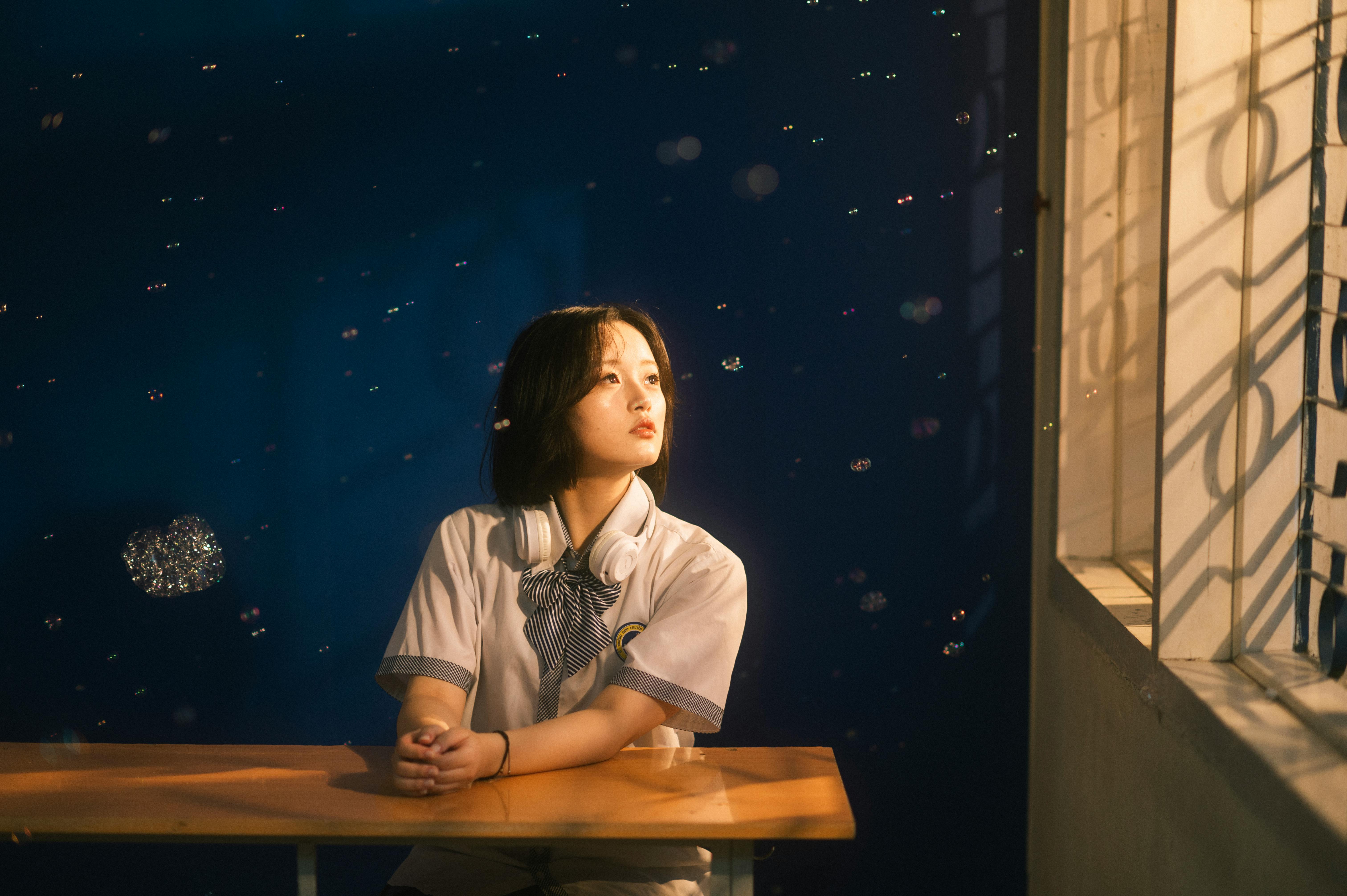 A student sits at a desk, gazing thoughtfully through a window, with gentle light and floating bubbles enhancing the serene atmosphere.