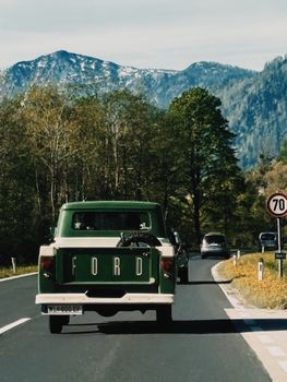 A vintage green truck driving on a mountain road surrounded by lush trees and snow-capped peaks.