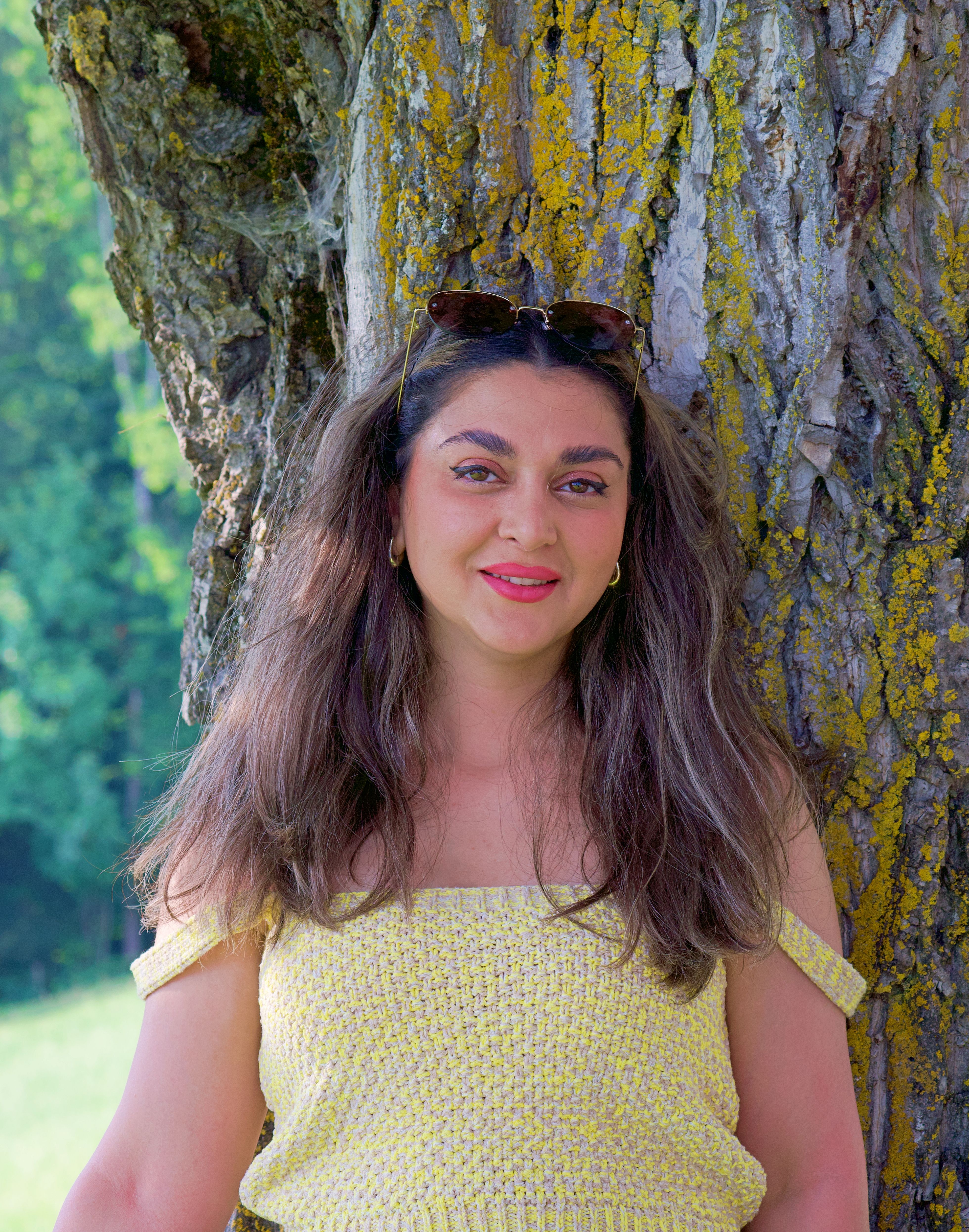 Smiling woman in a yellow dress posing by a tree in a sunny outdoor setting.