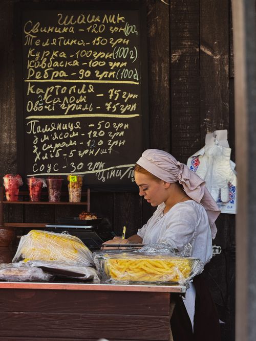 Free Woman in traditional clothing sells food at outdoor stall with prices listed in Cyrillic script. Stock Photo