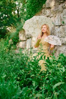 A woman in a vintage dress holds a parasol and fan, surrounded by lush greenery against a stone wall.
