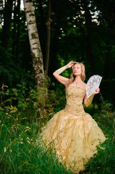 Woman in yellow ball gown with fan poised elegantly in a lush forest setting.