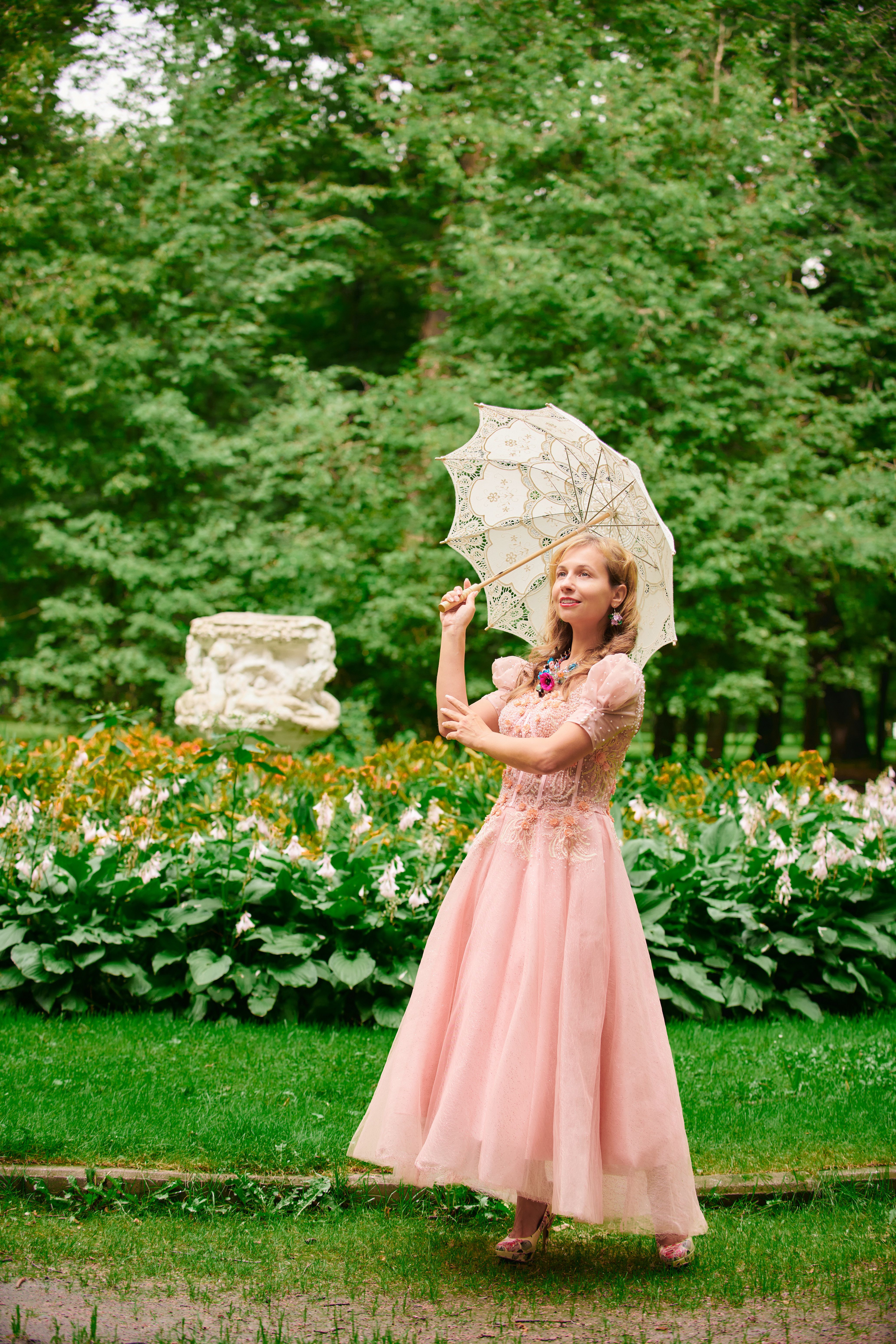 Elegant woman in a pink gown holding a parasol, standing in a lush garden.