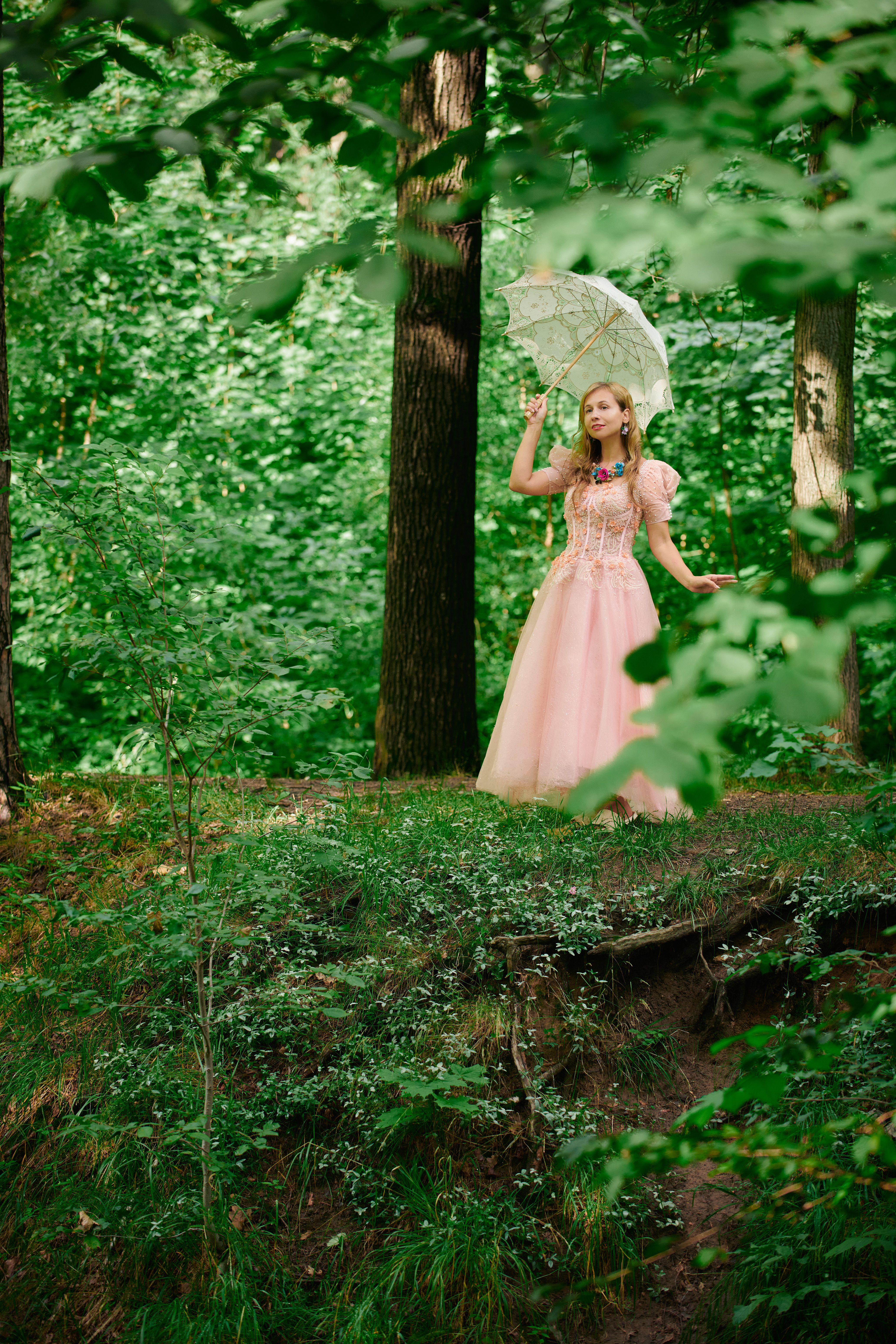 A woman in a pink gown with a lace parasol stands in a lush green forest, capturing a fairytale-like moment.