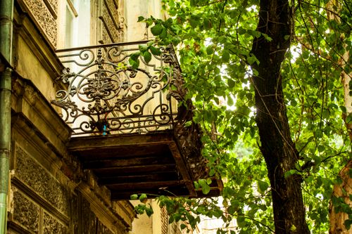 Free A decorative iron balcony with intricate design, nestled among lush green leaves on a sunny day. Stock Photo