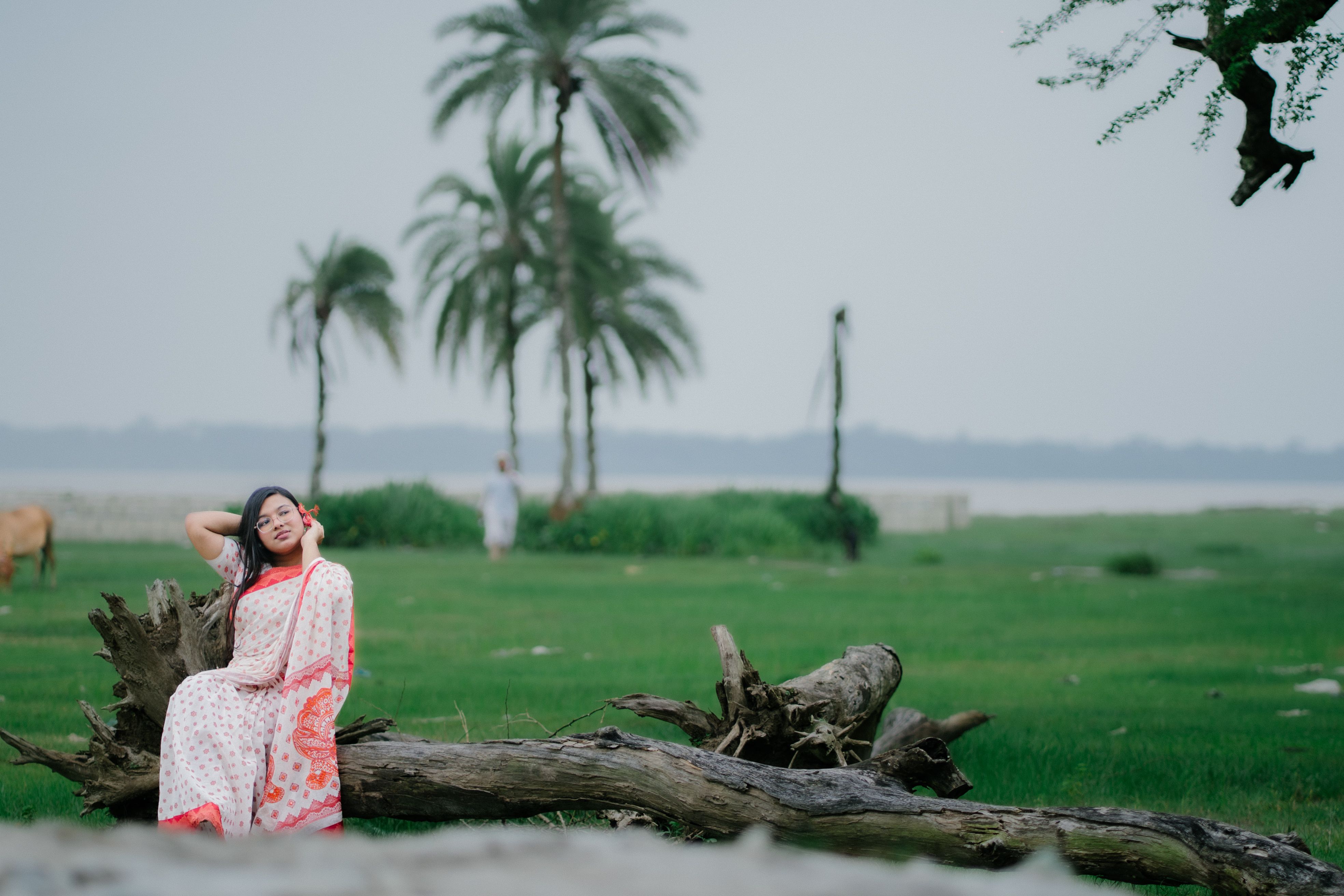 A woman in traditional attire enjoys a tranquil moment outdoors, surrounded by green grass and palm trees.