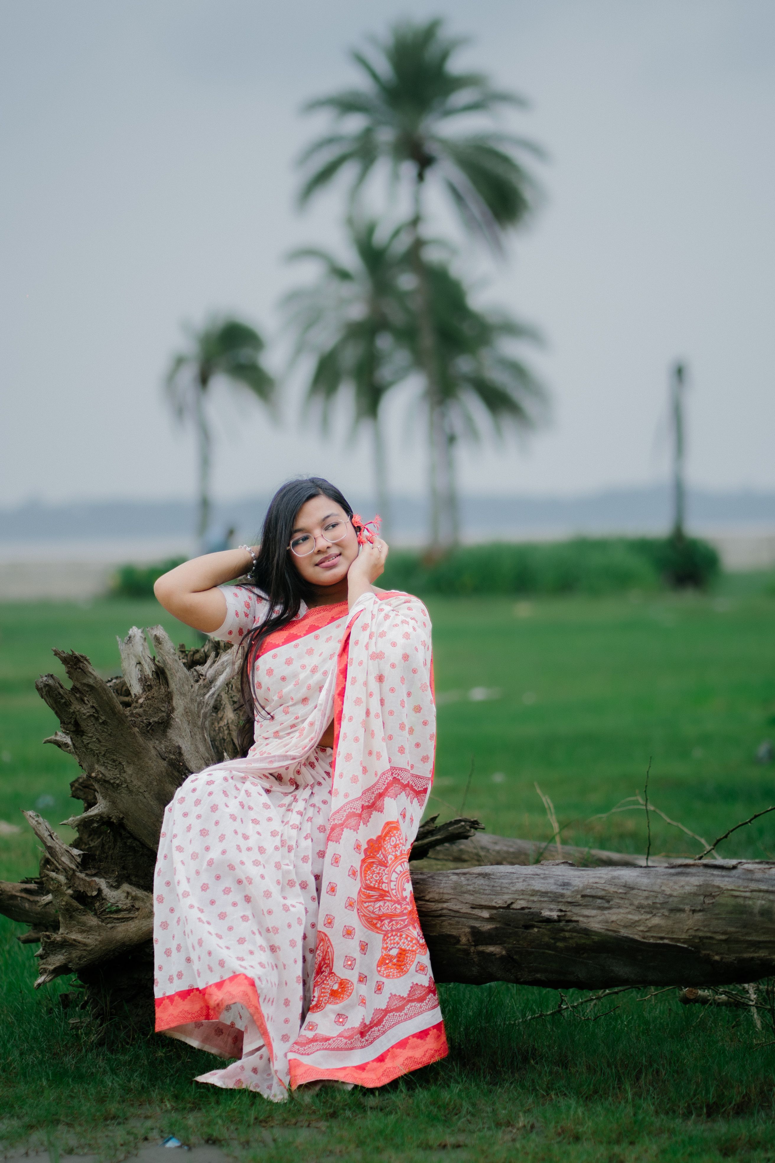 A young woman in a traditional saree sits on a log amidst a serene outdoor setting with palm trees.