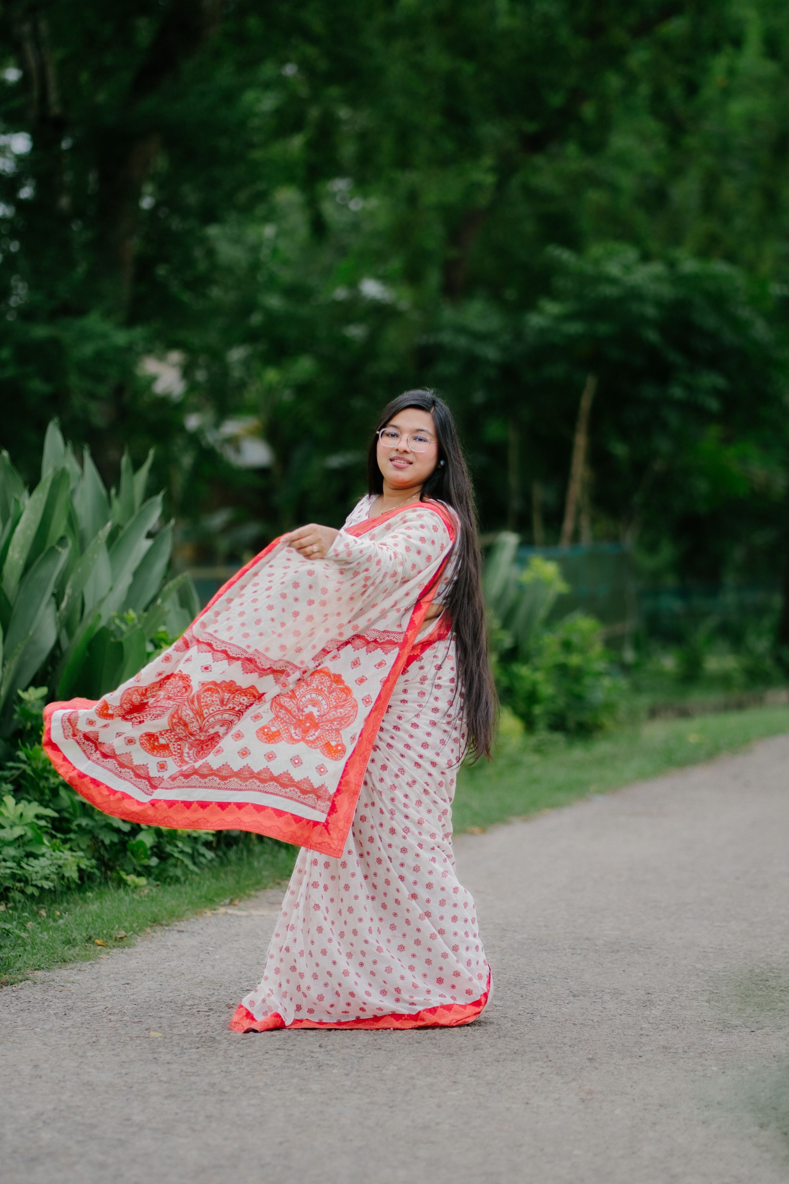 Woman in white saree with red floral design walking on a pathway surrounded by lush greenery.