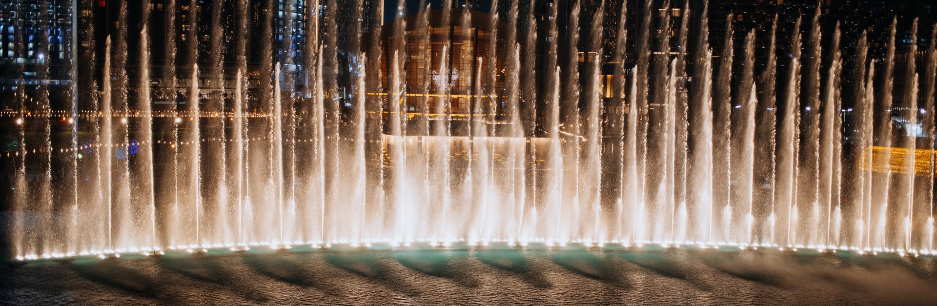 Illuminated water fountain show at night in downtown Dubai, surrounded by skyscrapers.