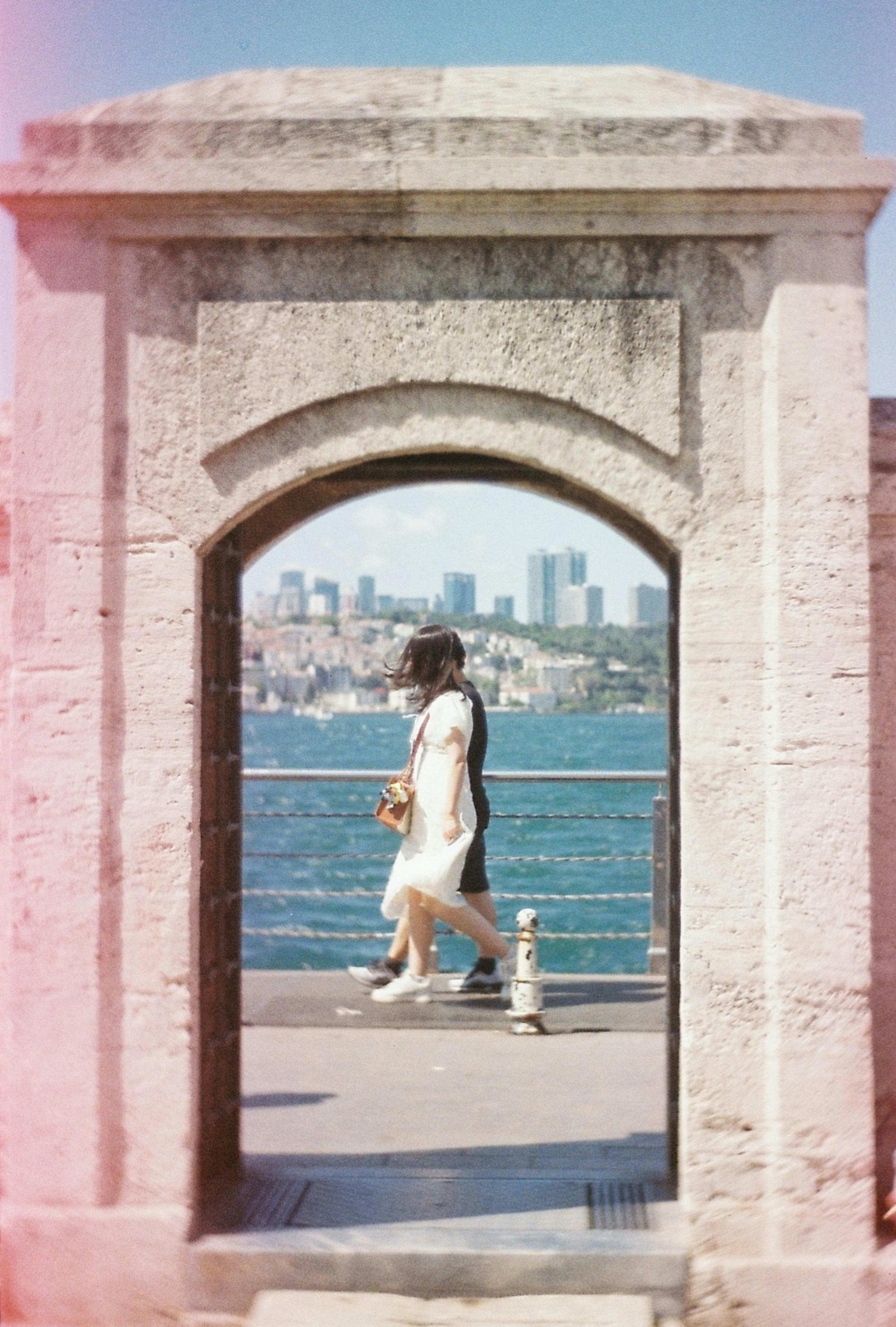 A scenic urban walk captured through a stone archway by the sea with a city skyline.