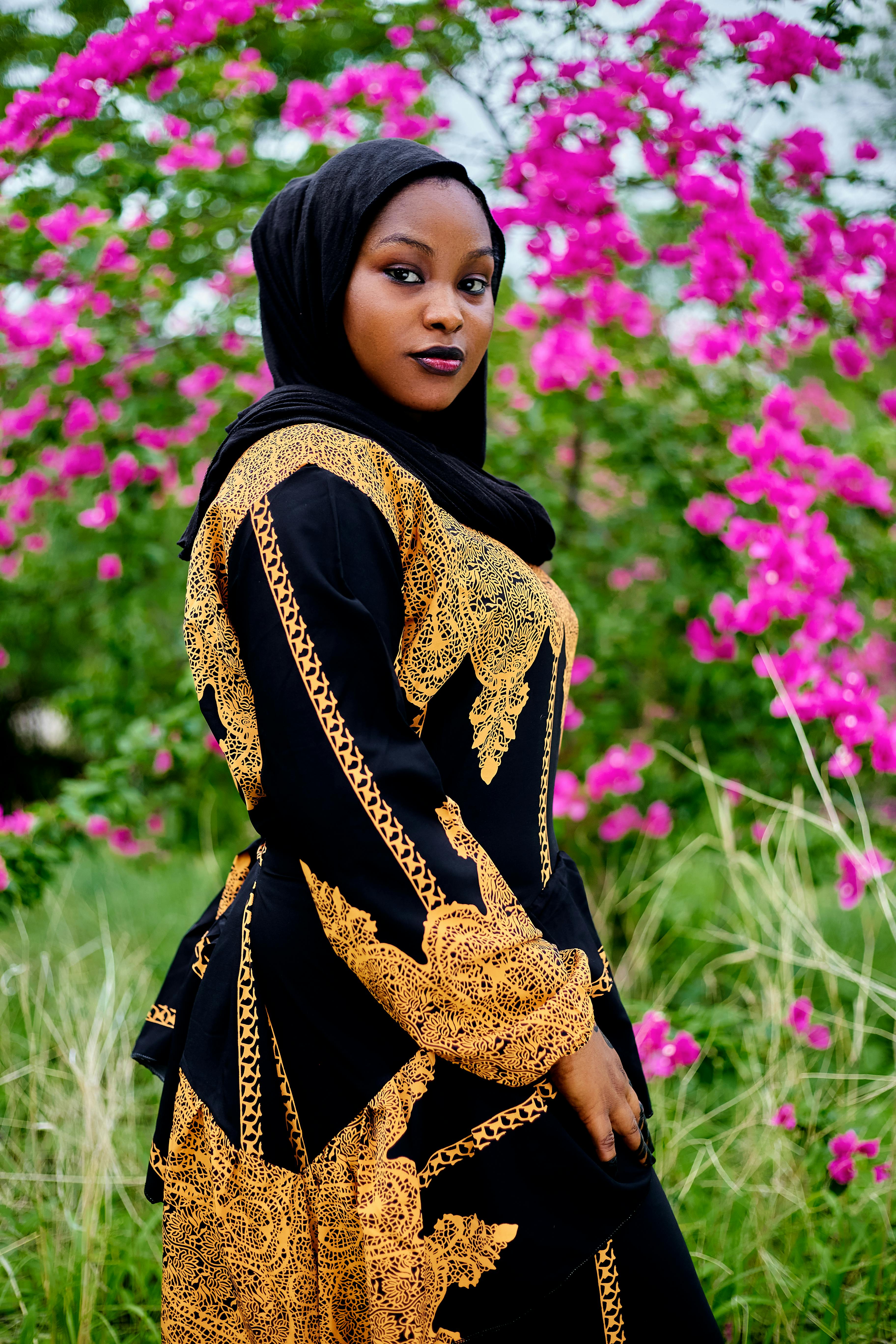 Elegant woman in traditional black and gold attire with vibrant pink flowers outdoors.