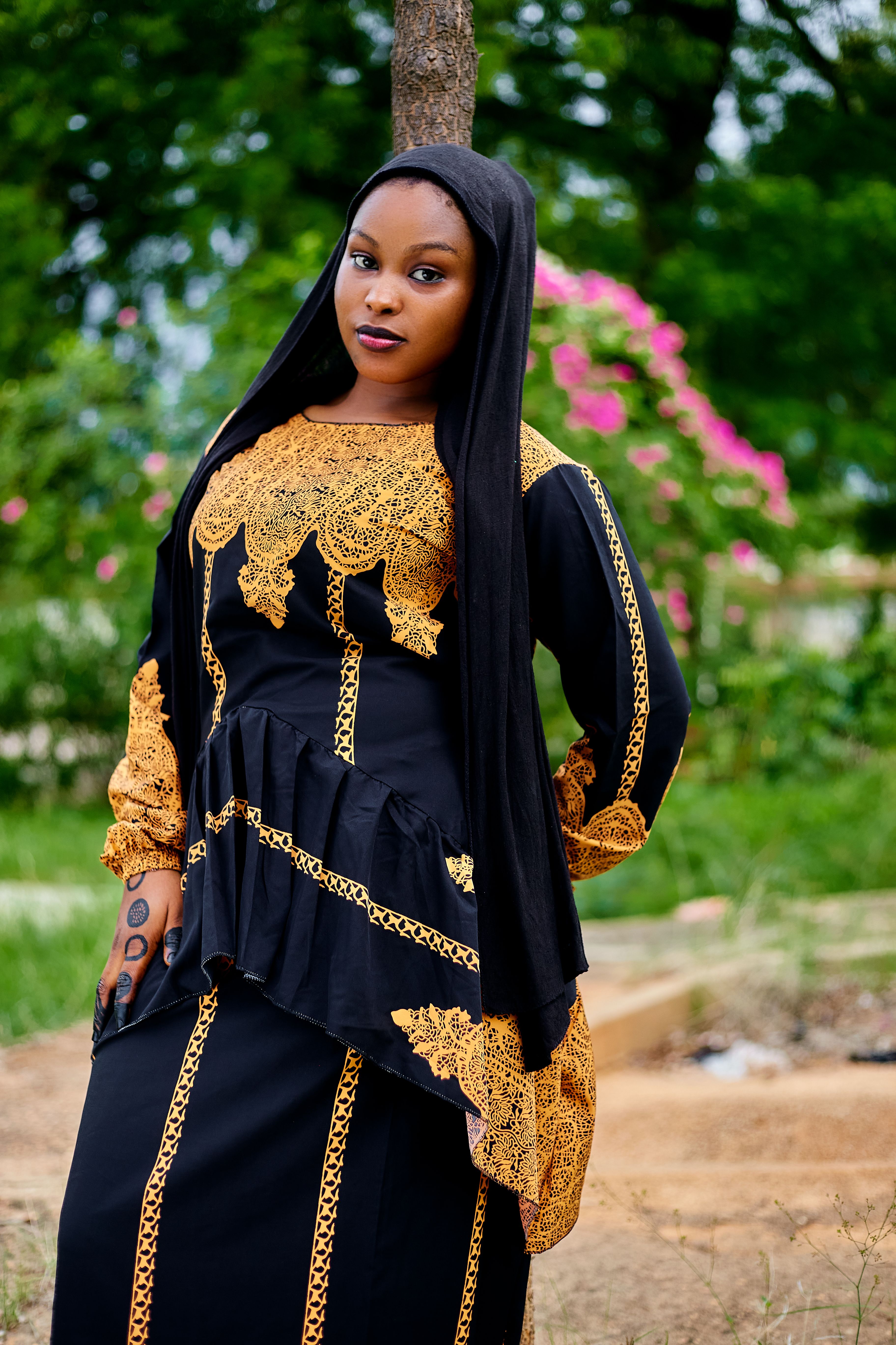 Woman in black and gold traditional attire standing outdoors surrounded by green foliage.
