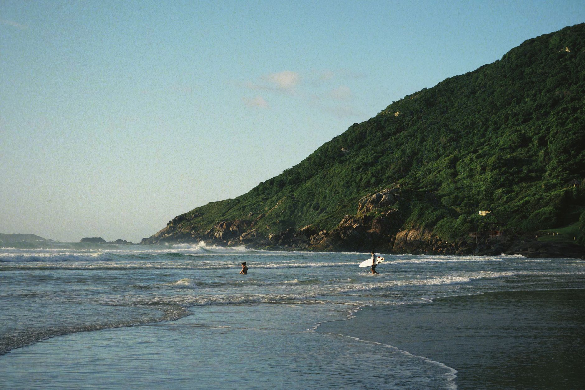 Peaceful summer day at Florianópolis beach with surfers and lush hills.