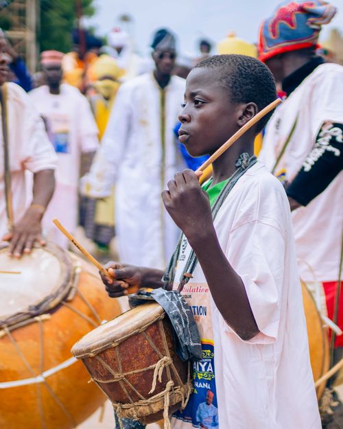 Free Teenager plays traditional drum at an outdoor cultural festival, showcasing vibrant clothing and lively atmosphere. Stock Photo