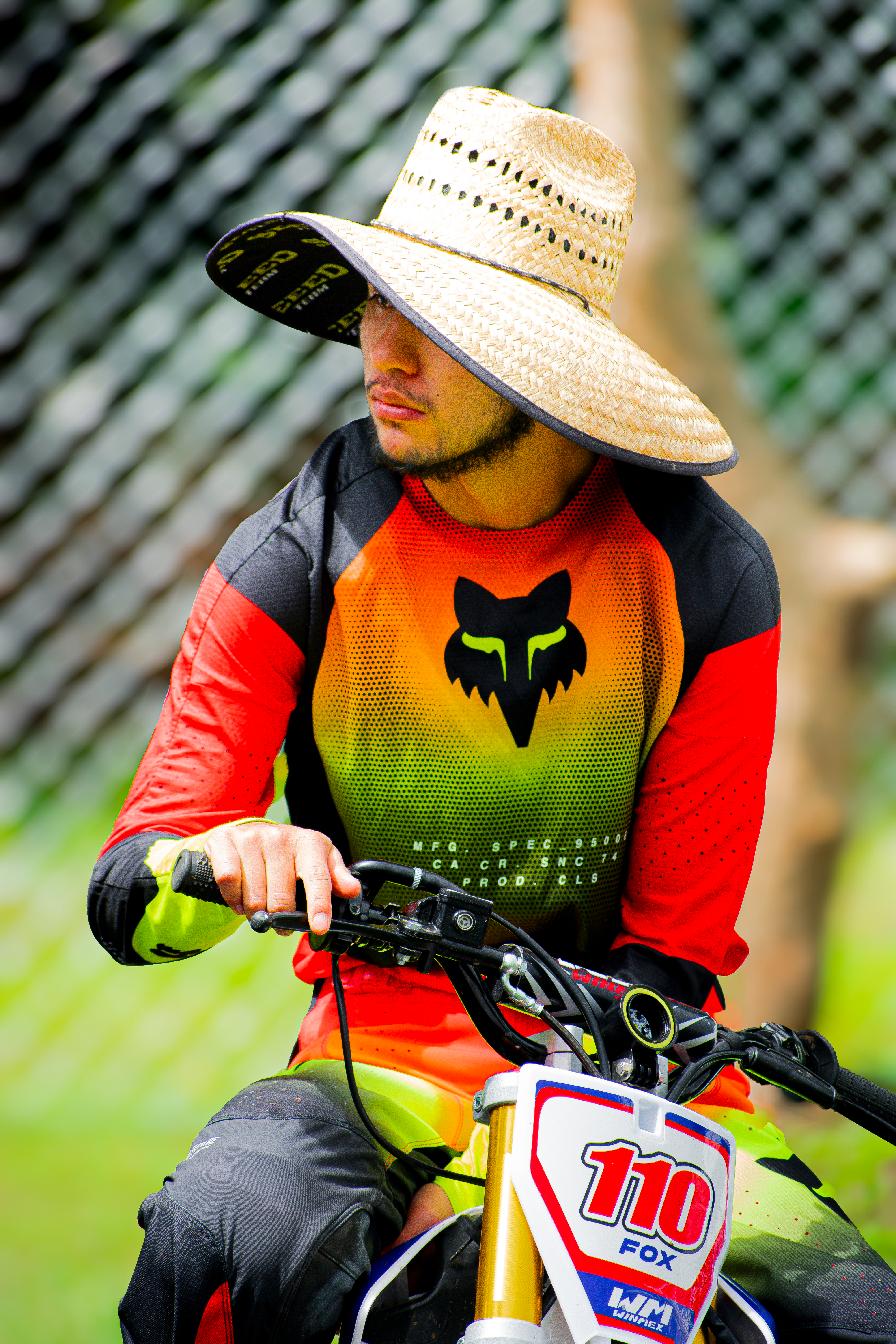 A young man wearing motocross gear and a straw hat rides a dirt bike outdoors in Mexico City.