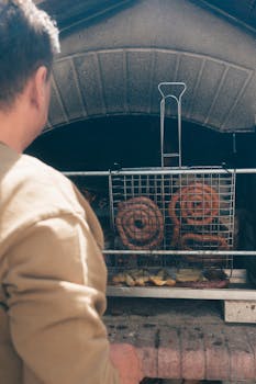 A person grills spiral sausages and vegetables in an outdoor oven, enjoying a sunny day.
