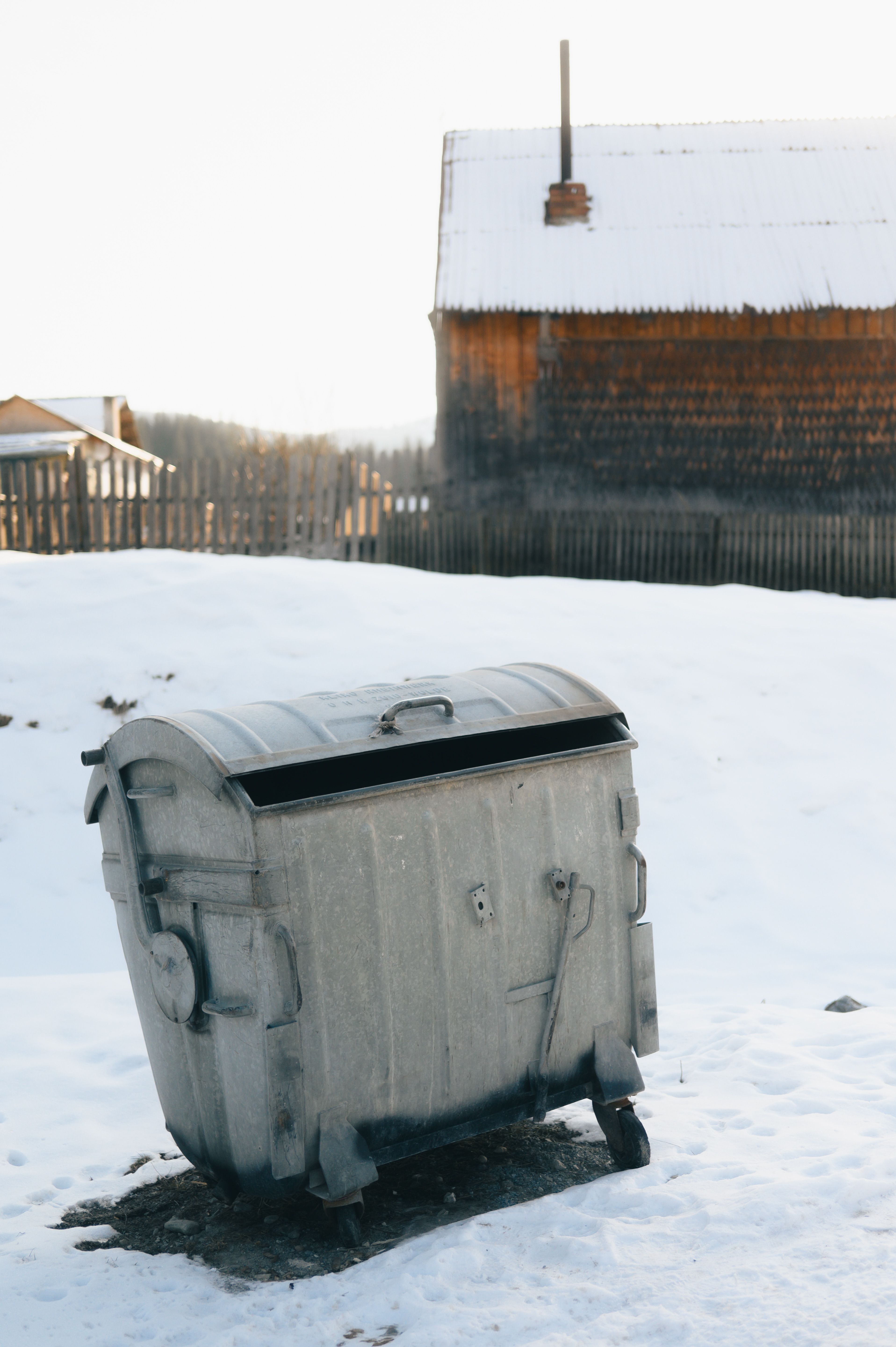 A vintage metal trash bin stands in a snowy rustic setting with wooden cabins.