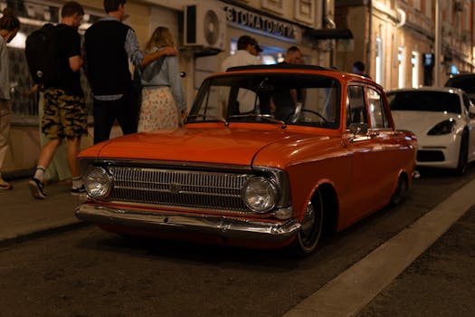 A classic orange car parked on a dimly lit city street at night, surrounded by people walking