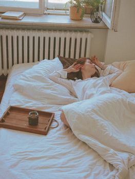 Woman relaxing in bed with morning sunlight, blanket, and coffee tray. Serenity captured.