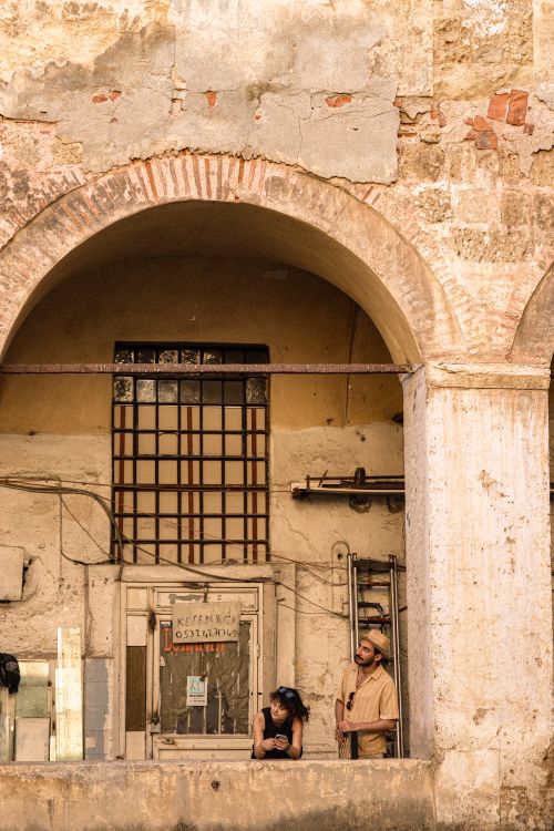 Free Two adults stand under a rustic archway observing an old building facade. Stock Photo