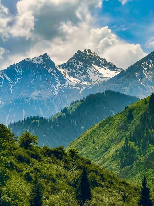 Free Scenic view of lush green hills and snowy mountains under a cloudy sky. Stock Photo