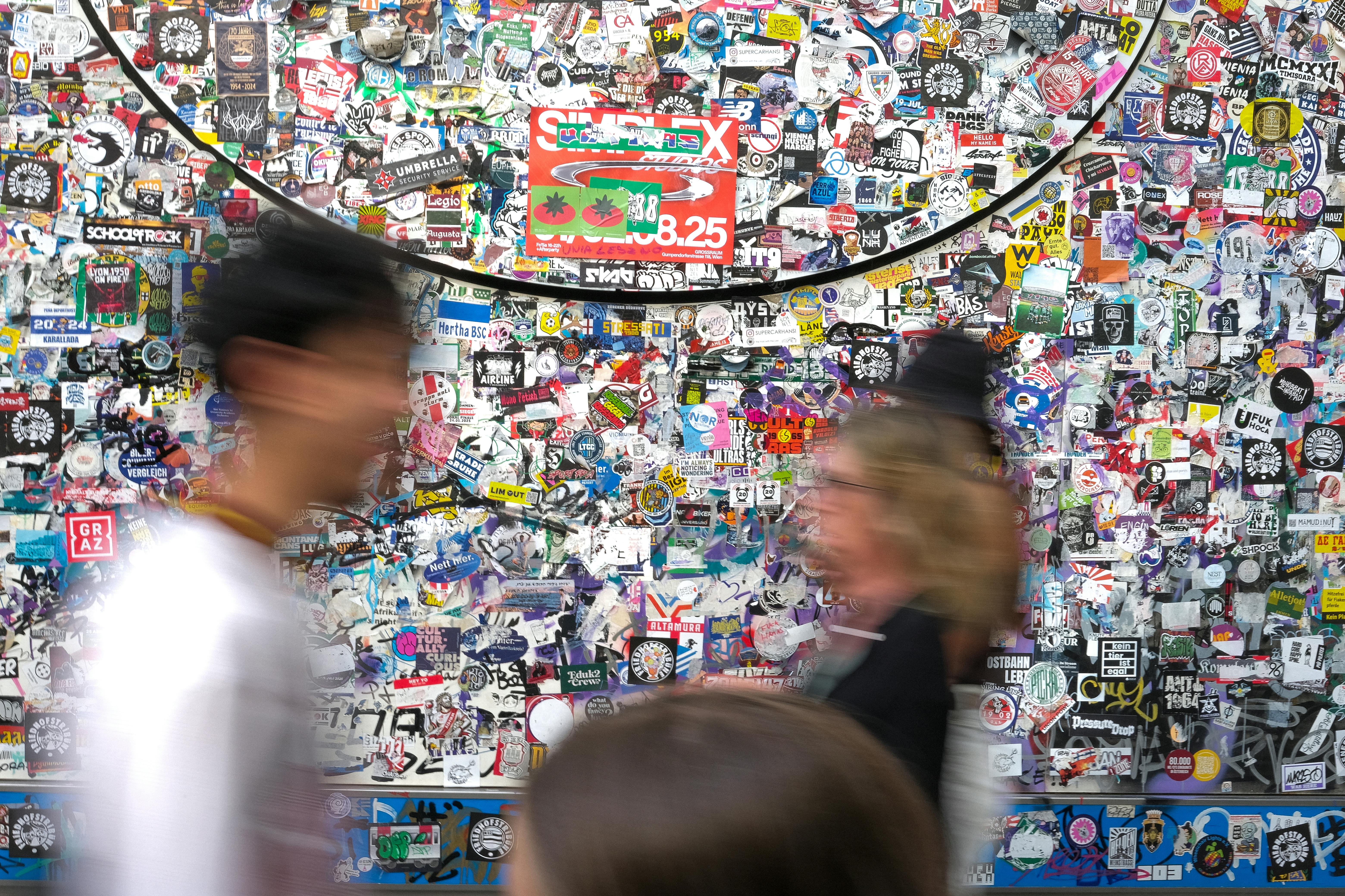 Dynamic urban scene with a colorful sticker-covered wall and blurred motion of people passing by, creating a lively street vibe.