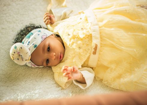 Cute baby in a yellow dress lying on a soft white blanket, wearing a headband. Warm and peaceful setting.