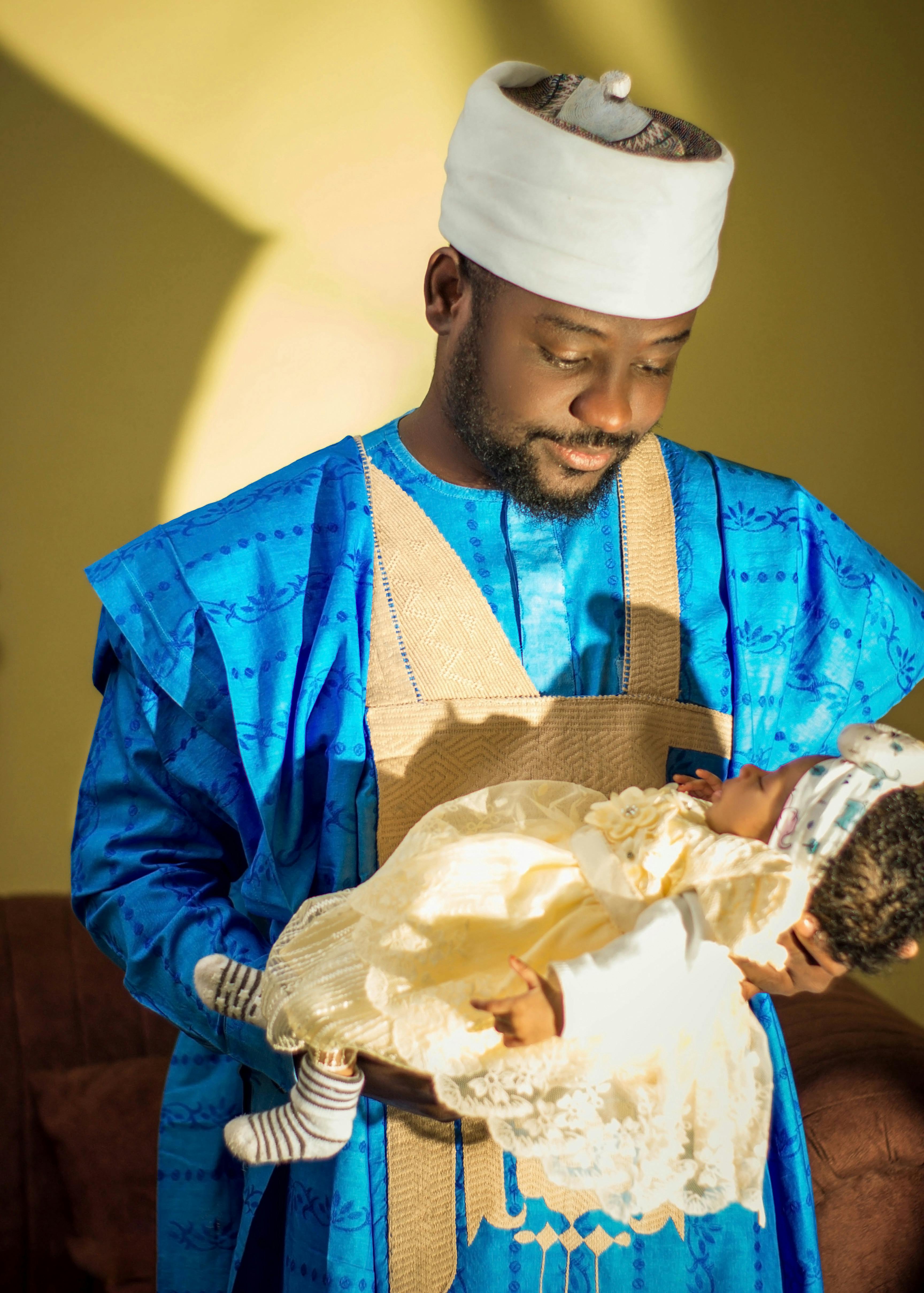 A father joyfully holds his newborn, dressed in traditional blue and white attire indoors.