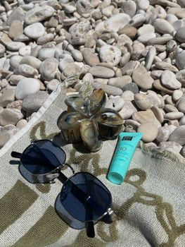 Flat lay of beach essentials including sunglasses and SPF cream on pebbles.