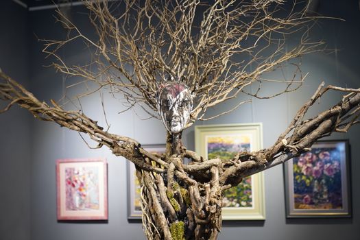 Artistic sculpture resembling a tree with a human face, displayed in a gallery setting.
