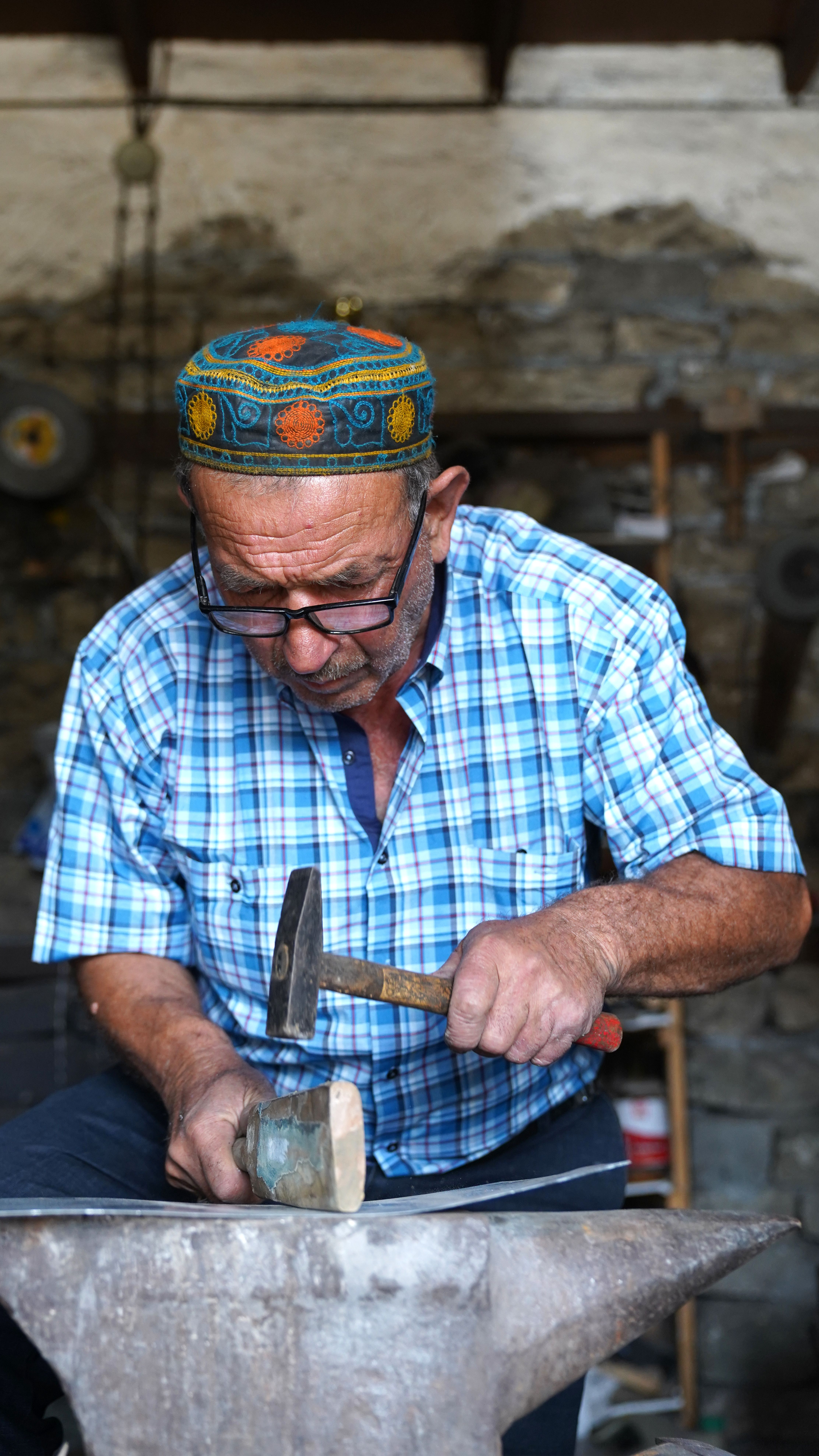 Elderly man working with hammer on metal in a rustic workshop setting.