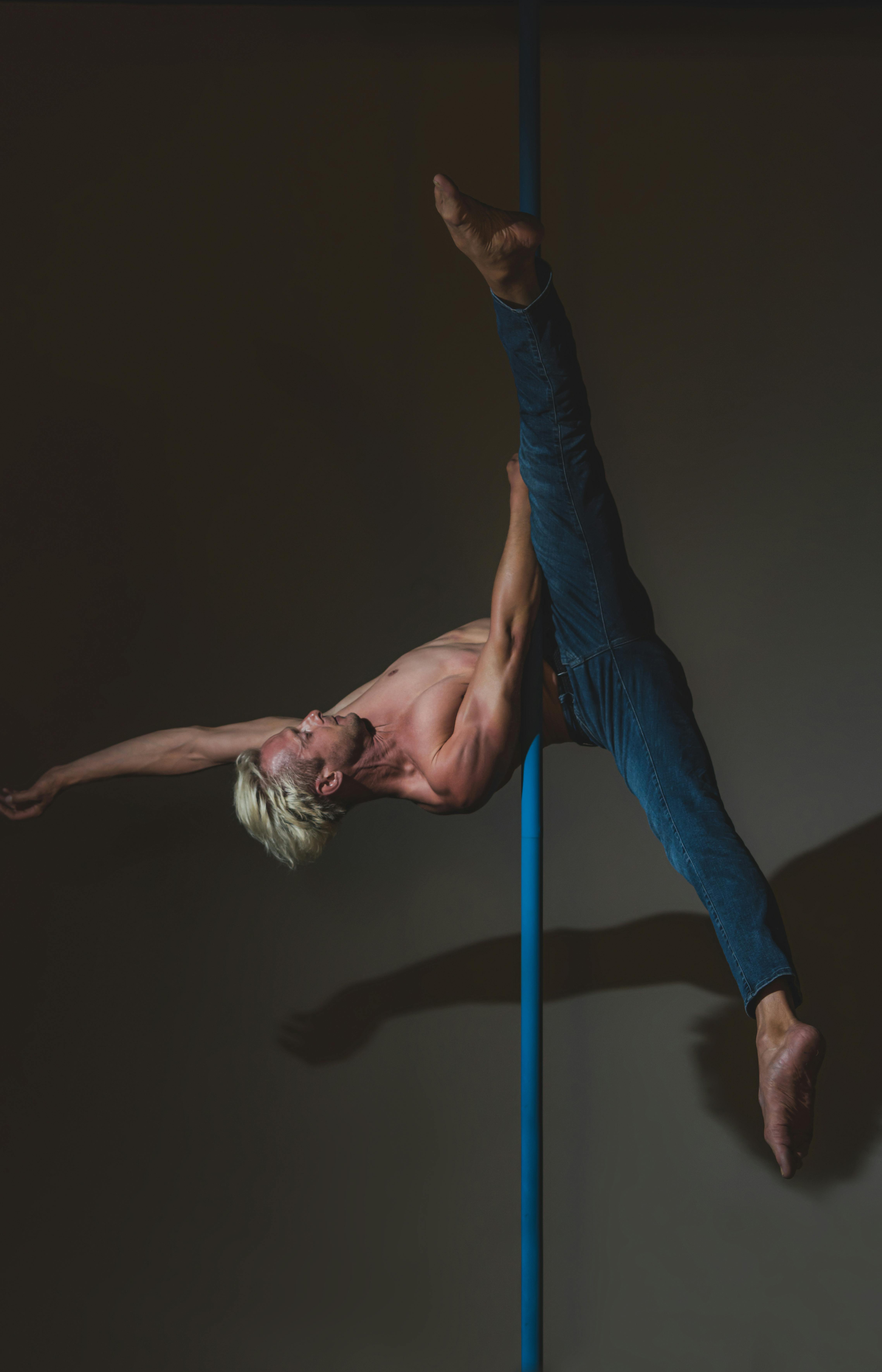 Danseur masculin exécutant une figure aérienne avancée de pole dance dans un studio professionnel sous éclairage dramatique.