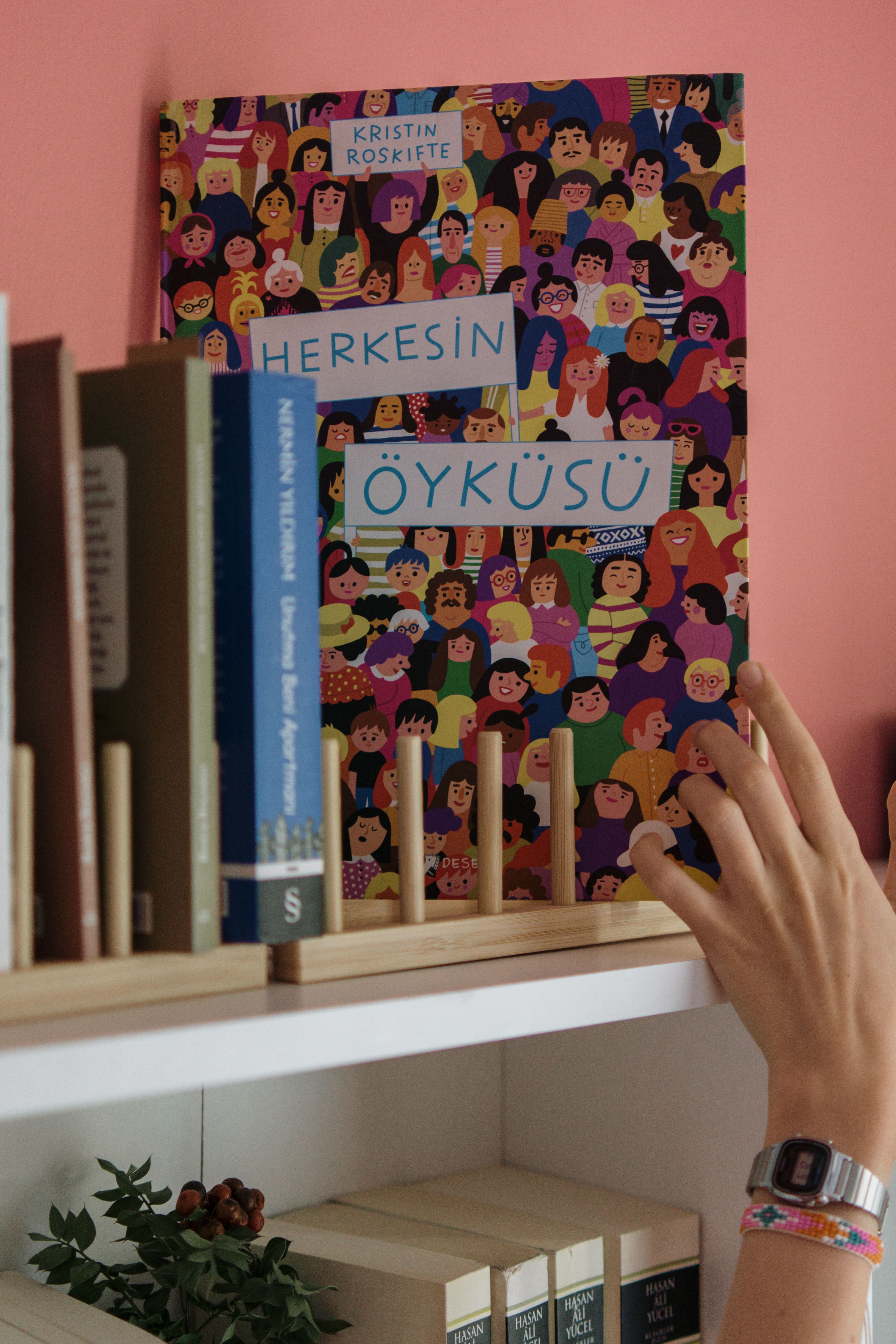 Hand reaching for a colorful illustrated book titled 'Herkesin Öyküsü' on a bookshelf.