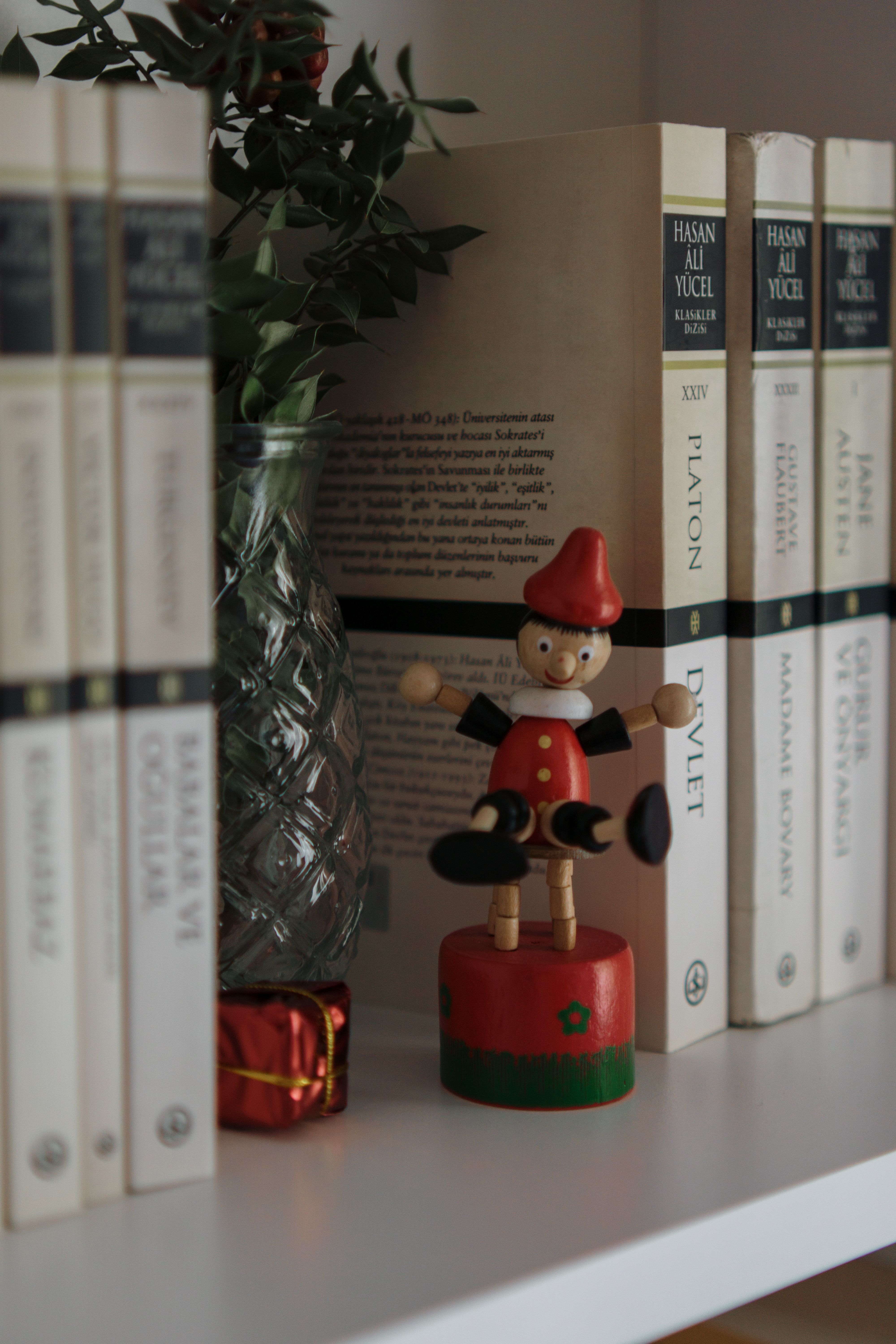 Simple bookshelf scene featuring a wooden puppet, classic books, and a glass vase.