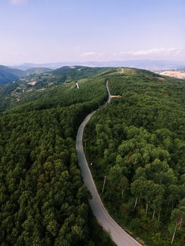 Aerial shot of a winding road through lush green forested hills under a clear sky.