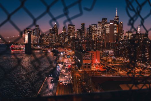 Captivating view of New York City skyline at night through a fence, featuring illuminated skyscrapers.