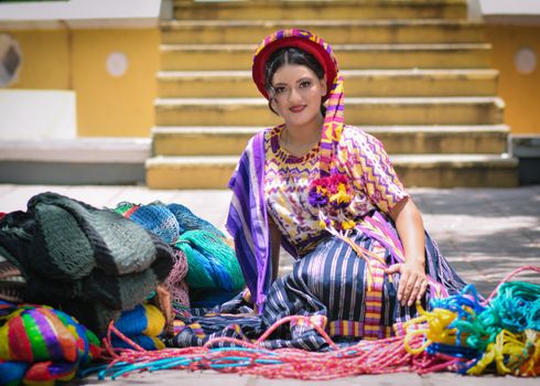 Woman in traditional colorful attire sitting with woven textiles, smiling outdoors.