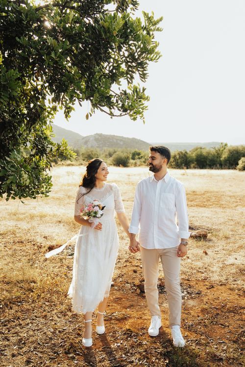 Free A couple in wedding attire walking hand in hand through a sunny outdoor landscape. Stock Photo